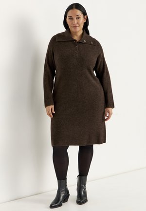 Vrouw draagt een bruine, gebreide jurk met lange mouwen en een geknoopte kraag, zwarte panty's en zwarte enkellaarsjes, staand tegen een effen witte achtergrond.