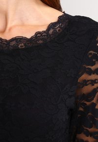Czarna koronkowa bluzka z kwiatowym wzorem, z karbowanym dekoltem i przezroczystymi długimi rękawami, podkreślająca fakturę i misternie wykonane detale.