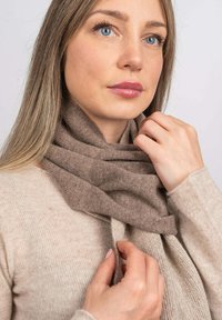 Sciarpa di lana marrone avvolta attorno al collo, con una texture morbida, su un maglione beige chiaro. Il modello ha i capelli lisci e la pelle chiara.
