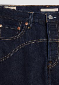Sötétkék farmernadrág réz szegecsekkel, arany varratokkal és jellegzetes ívelt varrási kialakítással. A 27-es méretű "ICON SKIRT" felirat található a címkén.