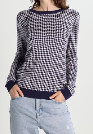 Frau trägt einen langärmeligen Pullover mit einem marineblauen und weißen Hahnentrittmuster, navyblaue Bündchen und Saum, kombiniert mit hellblauen Jeans und den Händen in den Taschen.