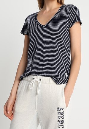 Navyblå og hvide stribede V-hals T-shirt med korte ærmer og løs pasform, med et lille logo på venstre side over kanten. Kombineret med lysegrå joggingbukser.