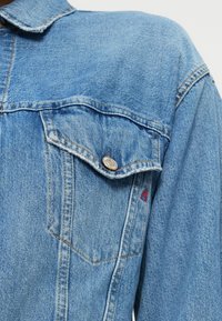 Τζάκετ τζιν σε ανοιχτό μπλε χρώμα με δύο μπροστινές τσέπες, μεταλλικές λεπτομέρειες στα κουμπιά, αντίθετες ραφές και ένα μικρό κόκκινο λογότυπο στην τσέπη.