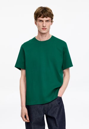 Junger Mann mit hellbraunen Haaren, der ein schlichtes grünes T-Shirt mit kurzen Ärmeln und dunkelblaue Jeans trägt, steht mit einer Hand in der Tasche.