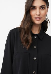 Vrouw met lang golvend bruin haar, dragend zilveren drop oorbellen en een zwarte met knopen gesloten blouse met zichtbare borstzakken tegen een witte achtergrond.