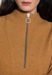 Brun, stickad tröja med hög krage och en silverfärgad dragkedja. Dragkedjan har en cirkulär dragflik. Texturen verkar mjuk och ribbad.