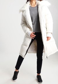 Femme portant un manteau long matelassé blanc avec une capuche en fourrure, un pull gris, un pantalon noir et des baskets noires, debout les jambes écartées.