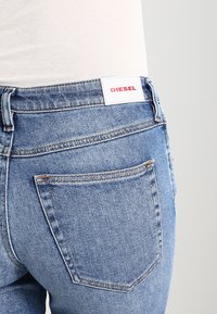 Blå jeans i denim med smal passform, med en vit etikett där det står "DIESEL" i rött. Detaljer inkluderar sömmar och en enda bakficka.