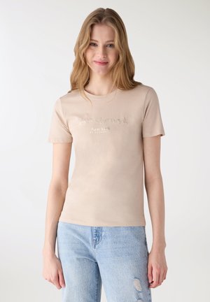 Giovane donna con capelli biondi lunghi che indossa una maglietta beige a maniche corte con la scritta "New York" e jeans azzurri strappati, in piedi.