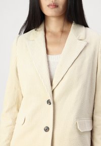 Kerman vaalean beige corduroy-takki, jossa on viistot laput, kaksi etutaskua ja yksittäinen nappikiinnitys. Ominaisuudet ovat pystysuunnassa riipputekstuuria ja erottuvia nappeja.
