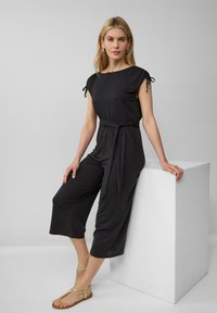 Schwarzer Jumpsuit mit kurzen Ärmeln, mit einem Band in der Taille und verkürzten Beinen. Verfügt über verstellbare Bindebänder an den Ärmeln. Getragen mit flachen Sandalen.