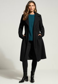 Schwarzer Wollmischungsmantel mit doppelreihigem Design, breiten Revers, seitlichen Taschen und langer Passform, kombiniert mit einem türkisfarbenen Pullover und schwarzen Jeans.