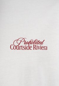 Texte rouge en cursive "Prohibited" au-dessus du texte avec empattement "Courtside Riviera" imprimé sur un fond blanc uni en tissu.