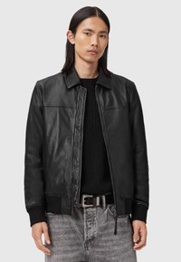 Ung man med rakt mörkt hår som bär en svart läderjacka över en svart tröja och urtvättade grå jeans med ett svart skärp.