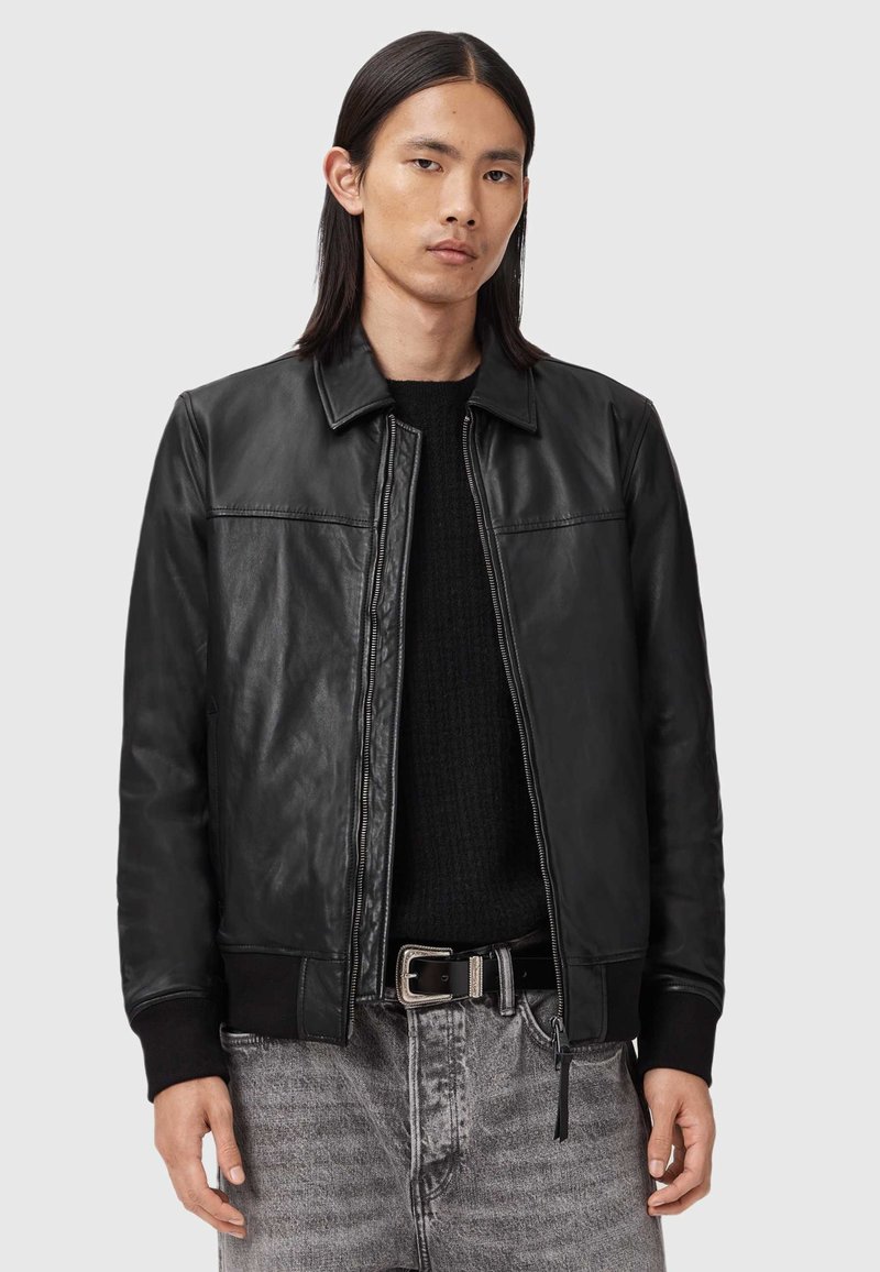 Ung man med rakt mörkt hår som bär en svart läderjacka över en svart tröja och urtvättade grå jeans med ett svart skärp.