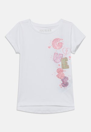Bílé tričko s krátkými rukávy s třpytivými růžovými, fialovými a zelenými písmeny, která svisle tvoří slovo "GUESS", obklopené malými růžovými srdíčky.