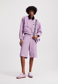 Lila kort jasje met twee voorzakken, gecombineerd met lila shorts. Witte en lila sneakers met lichtbruine zolen. Kleine lila handtas.