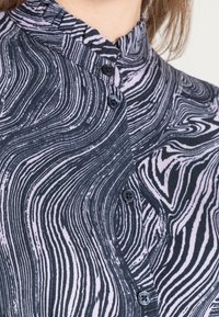 Napapaita, joka on varustettu musta-valkoisella aaltoilevalla kuviolla, röyhelökauluksella ja tummilla nappeilla edessä. Materiaali näyttää olevan tekstuuriltaan kevyt.