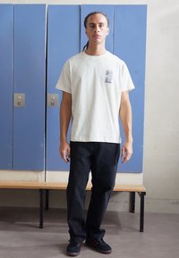 Weißes Baumwoll-T-Shirt mit aufgedrucktem Logo, schwarze Jeanshose und schwarze Sneaker, stehend vor blauen Schließfächern und einer Holzbank.