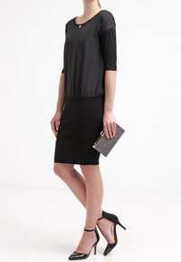 Schwarzes Kleid mit rundem Halsausschnitt und Dreiviertelärmeln; glatter Stoff oben, figurbetonter Rock. Schwarze Lackabsätze und eine silberne Clutch.