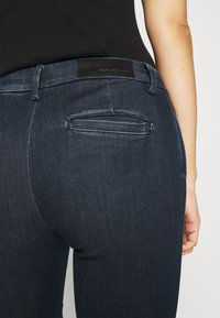 Jeans de color azul oscuro con un corte slim, fabricados en denim. Incluyen un bolsillo trasero y una etiqueta de marca de cuero negro en la cintura.