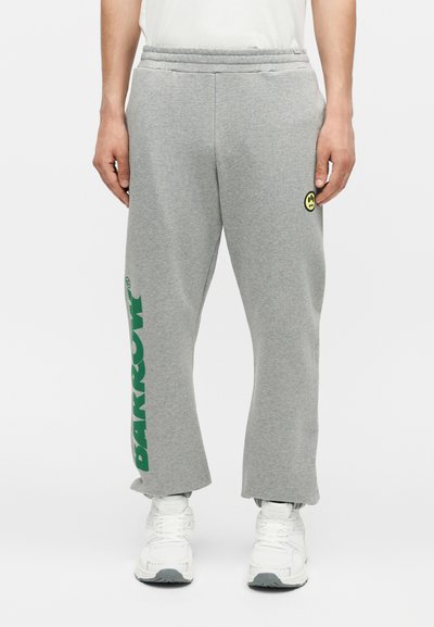 Άνδρας που φοράει γκρι φόρμες με πράσινο κείμενο "BARROW" στο αριστερό πόδι και ένα μικρό κίτρινο λογότυπο με χαμογελαστό πρόσωπο στο δεξί μηρό, σε συνδυασμό με λευκά αθλητικά παπούτσια.