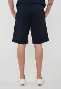 Pantaloni corti blu navy con una texture liscia, risvolti, due tasche posteriori e una vestibilità classica. Indossati con scarpe slip-on bianche in tela.