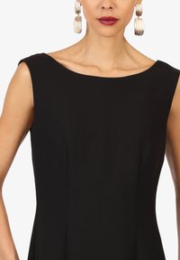 Robe sans manches noire avec un décolleté bateau. Tissu lisse avec un léger éclat. Grandes boucles d'oreilles circulaires en argent avec une texture striée.