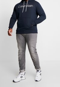 Felpa con cappuccio navy con logo bianco, tasca frontale; abbinata a jeans slim-fit grigi chiari e sneakers bianche con accenti navy.