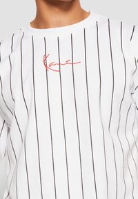 Biała koszula z cienkimi czarnymi pionowymi paskami i czerwonym kursywą napisem w okolicach kołnierza na górnej części ciała.