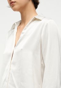 Γυναίκα που φοράει λευκή μπλούζα με υφή και μακριά μανίκια, με ανοιχτό γιακά, εμφανιζόμενη από τον λαιμό έως το πάνω μέρος του κορμού.