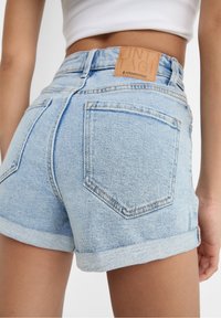 Pantalones cortos de mezclilla azul claro con dobladillo enrollado, usados por una persona con un top corto blanco, mostrando el bolsillo trasero y la etiqueta de marca marrón.