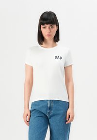 Donna con capelli neri lisci che indossa una maglietta bianca GAP e jeans blu su uno sfondo chiaro e semplice.