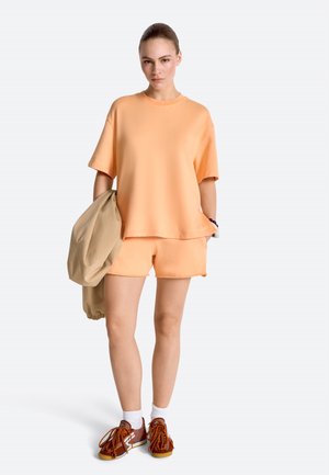 Giovane donna che indossa una maglietta oversize color pesca e pantaloncini, tiene in mano una giacca beige, indossa sneakers marroni con calzini bianchi, in piedi su sfondo bianco.