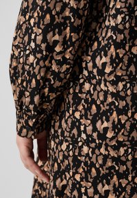 Μαύρο ύφασμα με μπεζ αφηρημένο ζωικό σχέδιο. Διαθέτει μανσέτα με κουμπιά και υφή στην επιφάνεια. Το μοτίβο ποικίλλει σε μέγεθος και σχήμα.