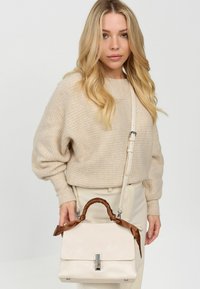 Beige handbag made of smooth material with a structured design, featuring a brown satin handle and a silver clasp. Adjustable strap included.