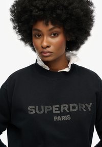 Zwarte sweatshirt met ribgebreide halslijn, voorzien van "SUPERDRY" en "PARIS" in studded letters. Zachte stof met een ontspannen pasvorm.