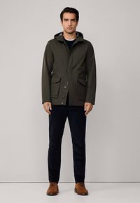 Olivovo zelená kapucňová bunda so zipsom a zapínaním na patentky, dvoma prednými vreckami a hladkou textúrou, nosená s tmavými nohavicami a hnedými topánkami.