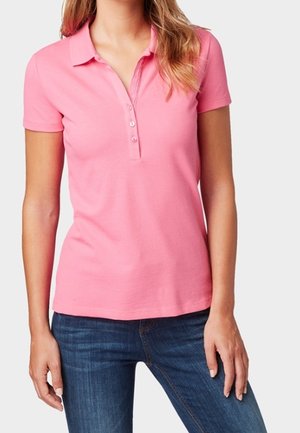 Kobieta w dopasowanej, różowej koszulce polo z krótkim rękawem i trzema guzikami, w ciemnoniebieskich dżinsach, stojąca na jednolitym tle.