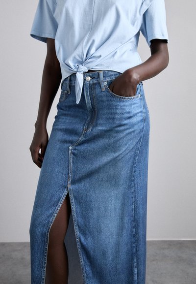 Jupe maxi en denim bleu moyen, avec une fente avant, un design décontracté et un style classique à cinq poches. Associée à un tee-shirt noué bleu clair.