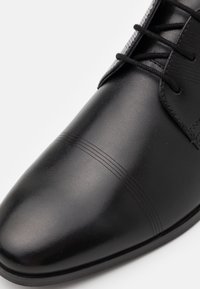 Chaussure habillée en cuir noir avec une surface lisse, un bout arrondi, des détails de couture horizontale subtils et des accents texturés près des lacets.