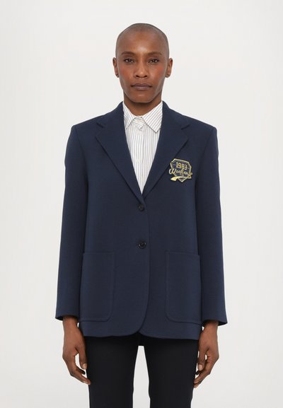 Námořnický oblekový blazer s dvěma předními kapsami, jedním knoflíkovým zapínáním, klopovými límci a zlatě vyšitým znakem s nápisem "1983 Weekend."