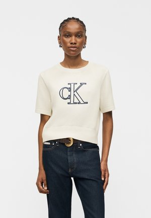 Kobieta z zaplecionymi włosami nosi biały sweter z krótkim rękawem z dużym logo „cK” oraz ciemne dżinsy z czarnym paskiem z złotą klamrą.