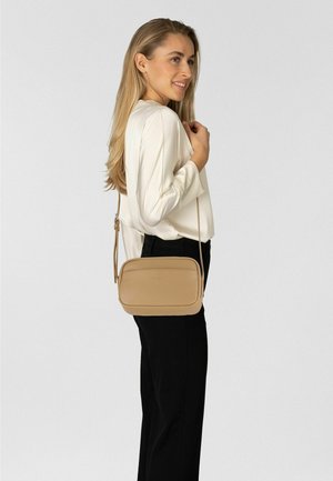 Beige leren crossbodytas met een gladde textuur, rechthoekige vorm, dunne riem en geperst logo. Model draagt een wit shirt en zwarte broek.