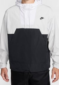 Kurtka typu pulower marki Nike, z czarną dolną częścią, białą górną częścią, regulowanym kapturem, przednią kieszenią i ściągaczem u dołu. Lekki materiał.