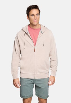 Hombre con sudadera con cremallera color beige claro sobre una camiseta roja deslavada y pantalones cortos verdes remangados, de pie sobre un fondo blanco.