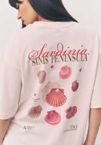 T-shirt oversize rose clair avec des motifs imprimés de coquillages et le texte « Péninsule de Sinis en Sardaigne » en rouge, accompagné de détails supplémentaires sur le musée et le lieu.