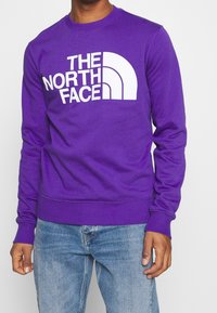 Fialová mikina s bílým textem „THE NORTH FACE“ a logem. Má kulatý výstřih, žebrované manžety a lem. Nositelná s modrými džínami.
