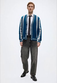 Μπλε ζακέτα με λευκές ρίγες, κλειστή με κουμπιά, φορεμένη πάνω από λευκό πουκάμισο και μαύρη γραβάτα. Γκρι τζιν με φαρδύ πόδι και γυαλιστερά μαύρα παπούτσια.