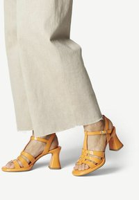 Orange patent leather sandals with a block heel, featuring multiple straps across the foot and a buckled ankle strap. Textured sole visible.